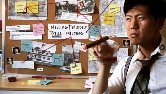 Man sits in front of cork board with series of photos of types of houses attached. Text printed in middle of cork board reads 